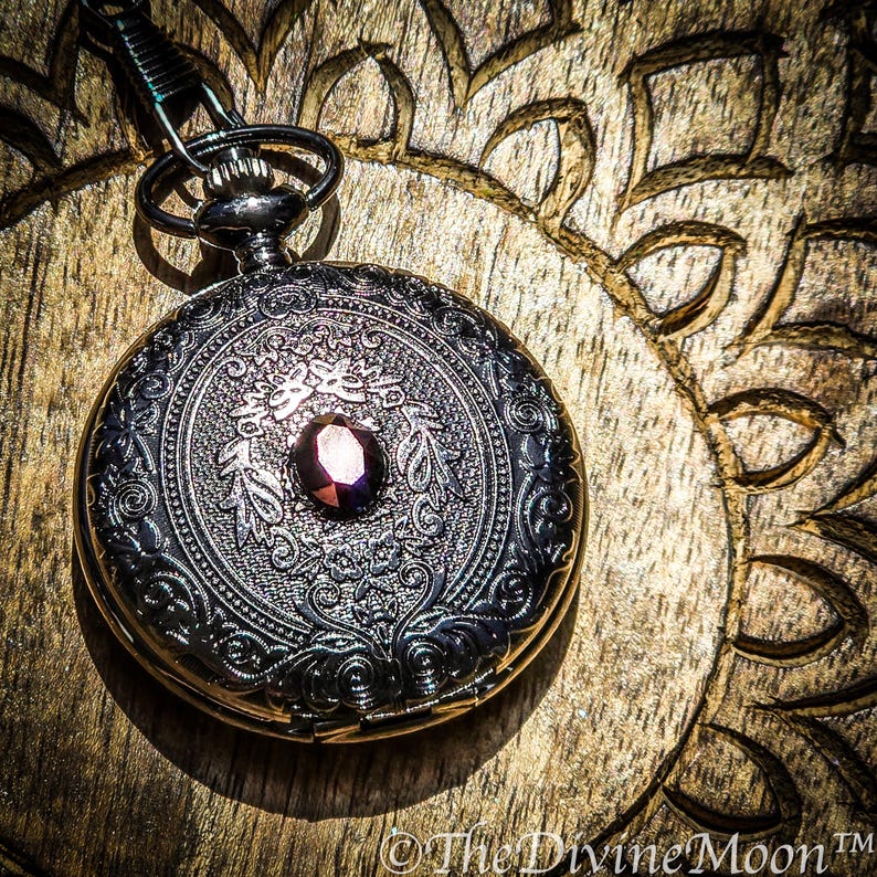 May include: A detailed, antique-style pocket watch with a dark metallic finish. The watch features an ornate, engraved design with a central, oval-shaped, dark red gemstone. The watch is suspended by a chain and rests on a wooden surface.