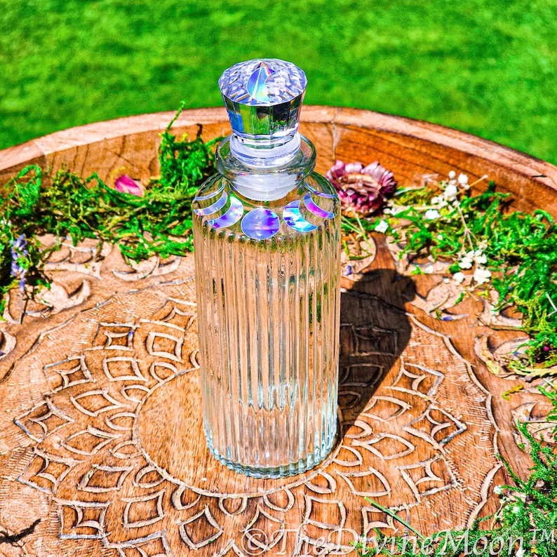 Puede incluir: Una botella de vidrio transparente con estr&iacute;as verticales y un tap&oacute;n facetado. El cuello de la botella presenta una serie de calcoman&iacute;as iridiscentes de las fases lunares. La botella tiene un cuerpo cil&iacute;ndrico y una tapa cristalina.
