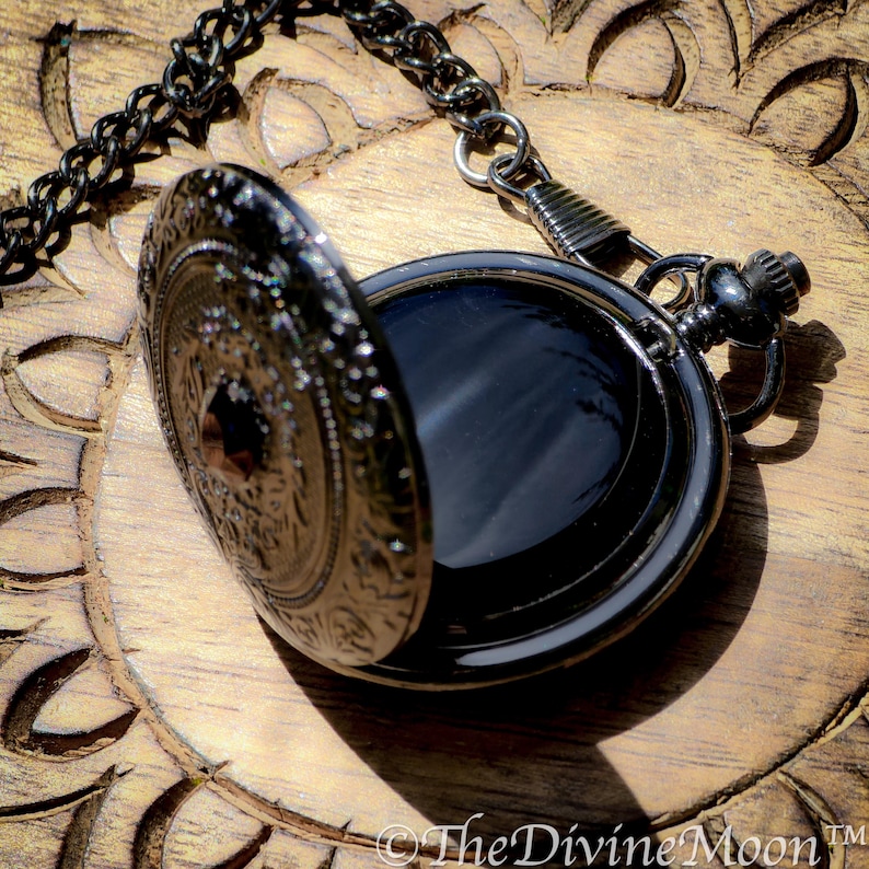 May include: An open, antique-style pocket watch with a black chain. The watch has an ornate, engraved design on the cover and a dark interior. The watch is resting on a wooden surface.