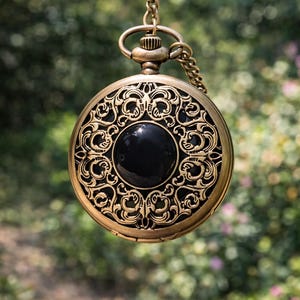 May include: Antique gold-colored pocket watch with an ornate, filigree design. The watch features a black, circular center and is suspended by a matching chain. The intricate details and vintage aesthetic make it a unique accessory.