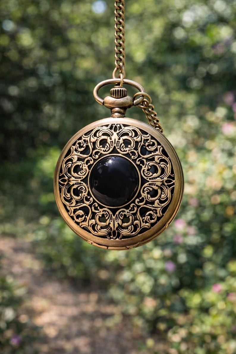 May include: Antique gold-colored pocket watch with an ornate, filigree design. The watch features a black, circular center and is suspended by a matching chain. The intricate details and vintage aesthetic make it a unique accessory.