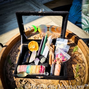 Puede incluir: Una caja de terciopelo negro llena de varios artículos para un ritual lunar. La caja contiene cristales, hierbas, velas y otros suministros mágicos. La caja está decorada con un diseño de luna y estrellas.