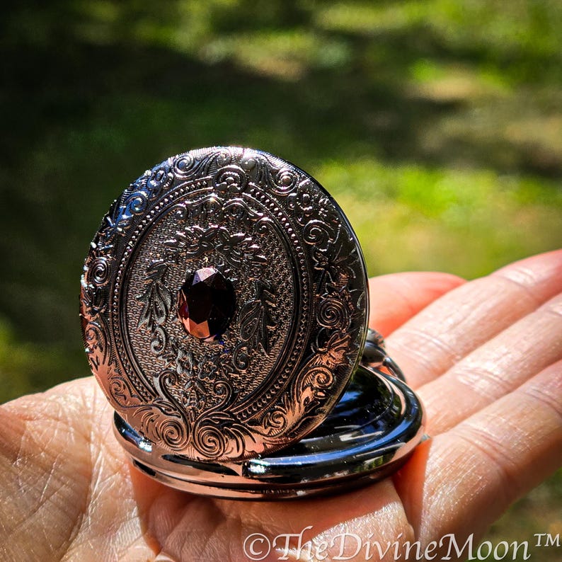 May include: An open, silver-colored, oval-shaped locket with intricate floral and scrollwork designs. A dark red gemstone is set in the center. The locket is held open, revealing the interior. The Divine Moon&trade; is printed at the bottom.