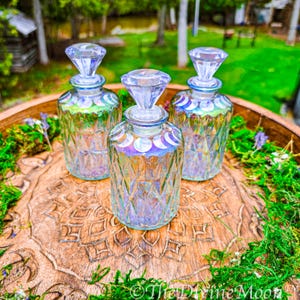 Puede incluir: Tres botellas de vidrio transparente con patrones de diamante y tapones facetados. Cada botella presenta una fila de calcoman&iacute;as blancas de las fases lunares alrededor del cuello. El vidrio tiene un acabado iridiscente.
