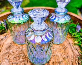 May include: Three clear glass bottles with diamond-shaped stoppers and iridescent moon phases designs. The bottles are sitting on a wooden surface.