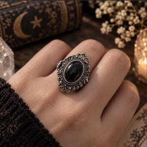 May include: A silver-toned metal locket ring featuring an oval-shaped black stone set in the centre. The ring band and setting are decorated with intricate, raised scrollwork and filigree detailing surrounding the smooth, dark central stone.