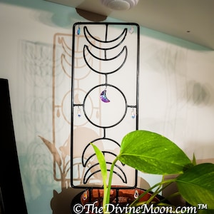 Könnte beinhalten: Schwarzer Metall-Wandbehang mit Mondphasen-Design, einem zentralen Kreis und Kristallakzenten. Ein kleiner, schillernder Mondanhänger hängt in der Mitte. Das The Divine Moon-Logo ist unten sichtbar.