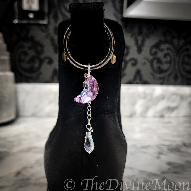 May include: A black boot with a silver charm attached to the top. The charm is a crescent moon-shaped crystal that is pink and iridescent. A smaller, teardrop-shaped crystal hangs from the moon charm.