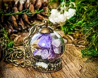 May include: A purple and white flower and amethyst crystal necklace pendant encased in a glass orb with a bronze chain and clasp. The pendant is on a wooden surface with green moss and white flowers.