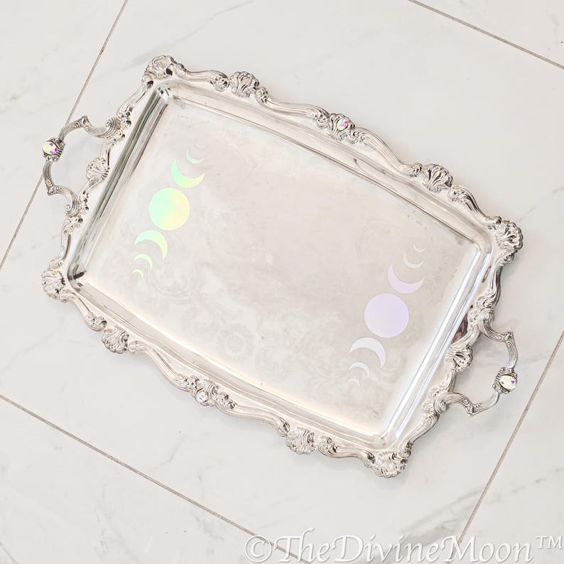 May include: A silver serving tray with ornate edges and two handles. The tray features a design of iridescent moon phases.