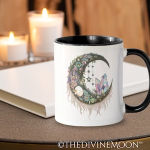 Puede incluir: Taza de cerámica blanca con borde y asa negros. La taza presenta un diseño de luna creciente con cristales, flores y estrellas. El texto "©THEDIVINEMOON™" está en la parte inferior de la taza.