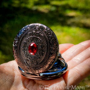 May include: Ornate silver-toned locket with a red gemstone center. The locket features detailed floral and scrollwork designs. The open locket reveals a polished interior. The Divine Moon&trade; is printed on the bottom right.
