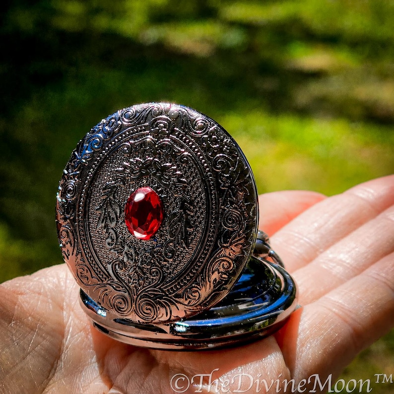 May include: Ornate silver-toned locket with a red gemstone center. The locket features detailed floral and scrollwork designs. The open locket reveals a polished interior. The Divine Moon&trade; is printed on the bottom right.