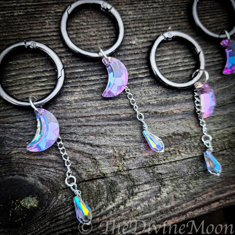 May include: Three silver keyrings with iridescent crescent moon charms and teardrop crystals hanging from them. The keyrings are on a wooden surface.