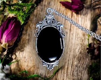 May include: A silver-toned pendant necklace featuring an oval-shaped black stone set within an ornate, filigree-style metal frame. The pendant hangs from a delicate silver chain, with the text The Divine Moon visible at the bottom.