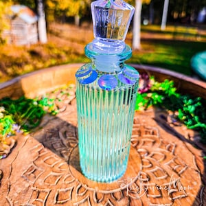 Puede incluir: Una botella de vidrio transparente con acanaladuras verticales y un tap&oacute;n facetado. El cuello de la botella presenta una serie de calcoman&iacute;as circulares iridiscentes que representan fases lunares en tonos azules, morados y plateados.