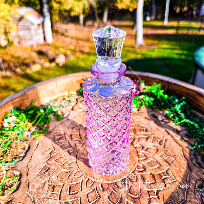 Puede incluir: Una botella de vidrio morado con textura de patr&oacute;n de diamantes y un tap&oacute;n transparente facetado. El cuello de la botella est&aacute; decorado con una serie de s&iacute;mbolos blancos de fases lunares. La botella tiene un cuerpo cil&iacute;ndrico.