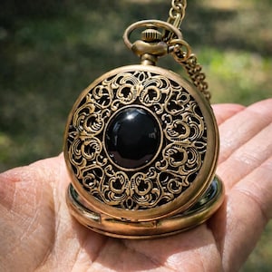 May include: Antique gold-colored pocket watch with an ornate, filigree design. The watch features a large, black, circular stone in the center. A matching chain is attached to the top. The watch is open, revealing the inner workings.