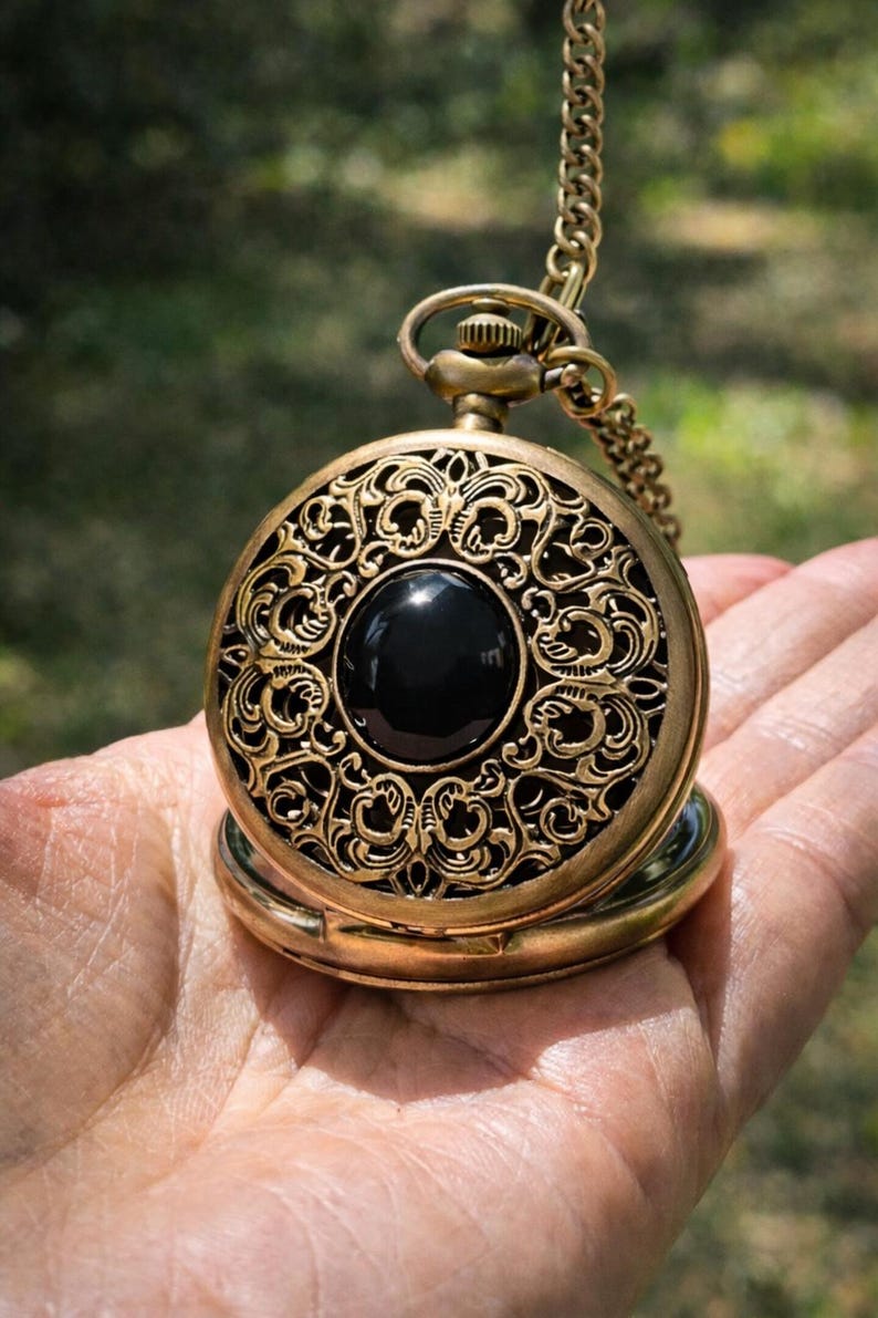 May include: Antique gold-colored pocket watch with an ornate, filigree design. The watch features a large, black, circular stone in the center. A matching chain is attached to the top. The watch is open, revealing the inner workings.