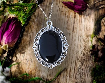 May include: A silver necklace with a black oval pendant in an ornate silver frame. The pendant is set against a wooden background.