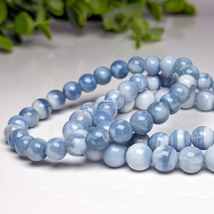 May include: A beaded bracelet featuring smooth, round, polished stones in varying shades of light blue, slate blue, and white. The stones are strung together in a continuous loop, displaying a marbled, semi-translucent appearance with a glossy finish.
