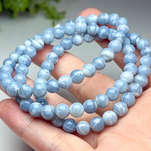 May include: A beaded bracelet featuring smooth, round, light blue stones with subtle white marbled patterns. The beads are uniform in size and strung together on an elastic cord, creating a continuous circular loop of polished, sky-colored spheres.