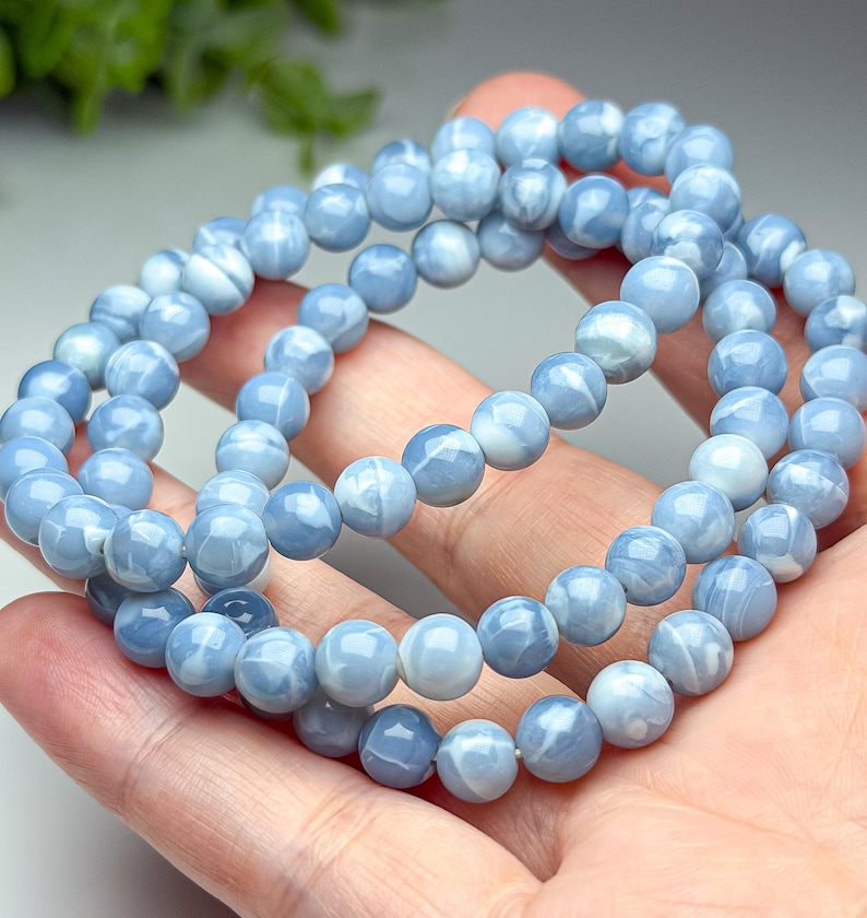 May include: A beaded bracelet featuring smooth, round, light blue stones with subtle white marbled patterns. The beads are uniform in size and strung together on an elastic cord, creating a continuous circular loop of polished, sky-colored spheres.