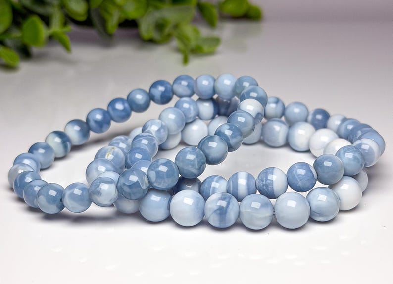 May include: A beaded bracelet featuring smooth, round, polished stones in varying shades of light blue, slate blue, and white. The stones are strung together in a continuous loop, displaying a marbled, semi-translucent appearance with a glossy finish.