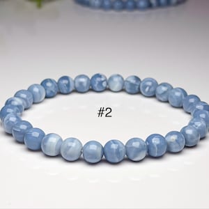May include: A stretch bracelet composed of smooth, round, light blue beads with subtle white veining and a glossy finish. The bracelet is marked with the text #2 in the center, indicating a specific item in a collection of beaded jewelry.