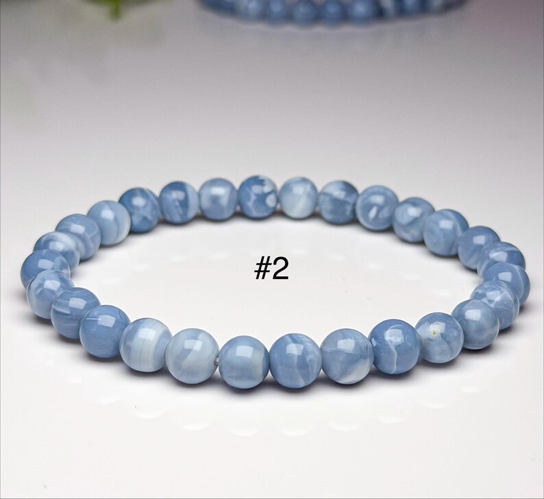 May include: A stretch bracelet composed of smooth, round, light blue beads with subtle white veining and a glossy finish. The bracelet is marked with the text #2 in the center, indicating a specific item in a collection of beaded jewelry.