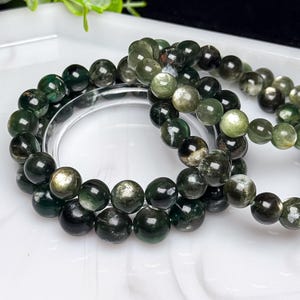 May include: A beaded bracelet featuring polished, spherical dark green and black gemstones with visible internal inclusions and metallic, reflective patches. The beads are strung together in a circular loop, displaying varying shades of deep forest green.