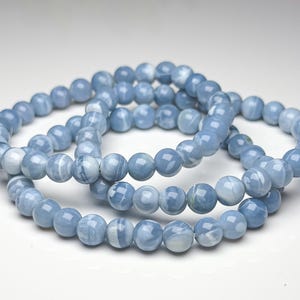 May include: A beaded bracelet featuring smooth, round, light blue stones with white marbled patterns. The beads are uniform in size and strung together in a circular loop, displaying a glossy, polished finish throughout the entire piece.