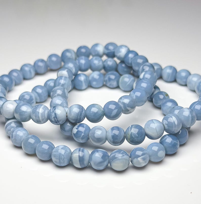May include: A beaded bracelet featuring smooth, round, light blue stones with white marbled patterns. The beads are uniform in size and strung together in a circular loop, displaying a glossy, polished finish throughout the entire piece.