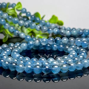May include: Strands of blue aquamarine gemstone beads. The beads are round and have a translucent quality, with varying shades of blue. The beads are arranged in a circular pattern, with a few strands in the background.