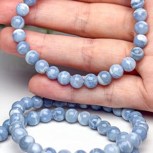 May include: A stretch bracelet composed of smooth, round, light blue beads with white marbled patterns. The beads are uniform in size and strung together on an elastic cord, creating a circular shape that is flexible and easy to wear on the wrist.