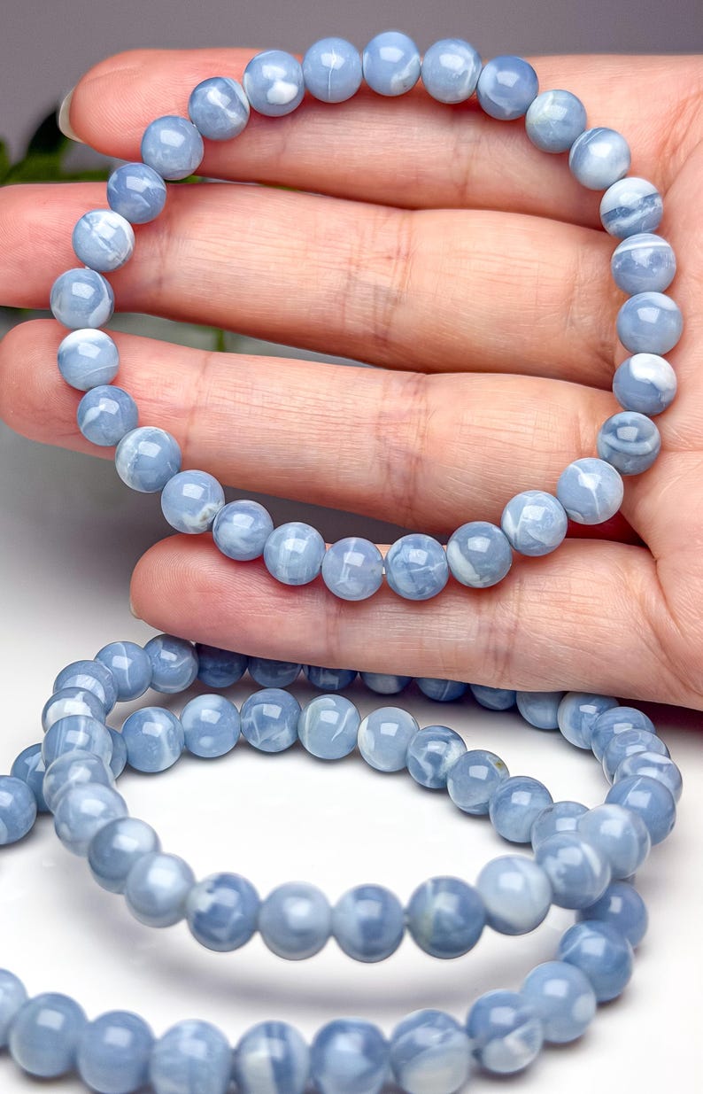 May include: A stretch bracelet composed of smooth, round, light blue beads with white marbled patterns. The beads are uniform in size and strung together on an elastic cord, creating a circular shape that is flexible and easy to wear on the wrist.