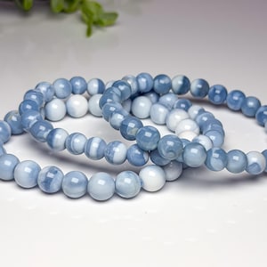 May include: Three stretch bracelets composed of smooth, round beads in varying shades of light blue, medium blue, and opaque white. The beads feature a polished finish and are arranged in a circular pattern, creating a cohesive set of jewelry.