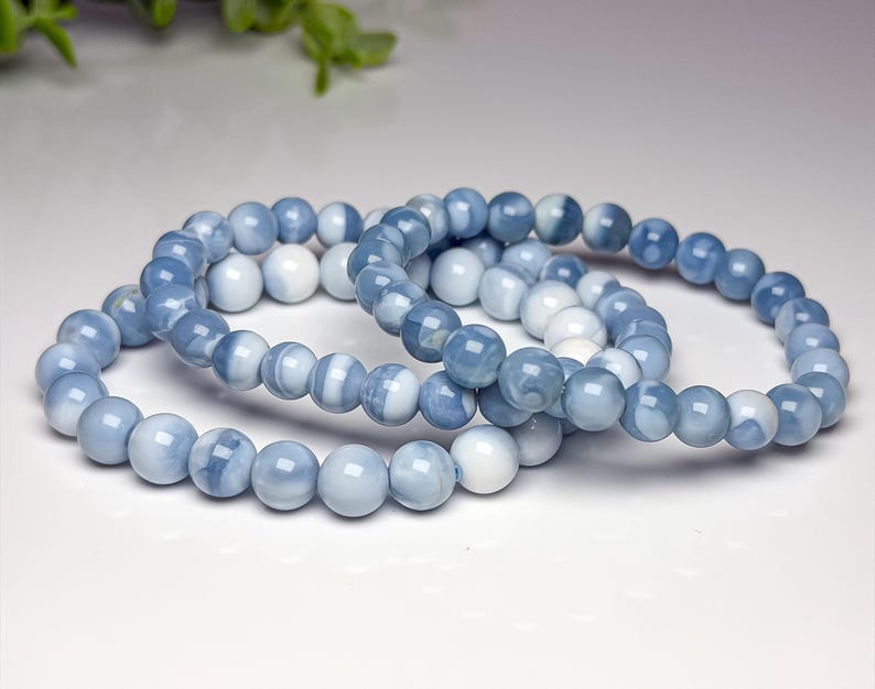 May include: Three stretch bracelets composed of smooth, round beads in varying shades of light blue, medium blue, and opaque white. The beads feature a polished finish and are arranged in a circular pattern, creating a cohesive set of jewelry.