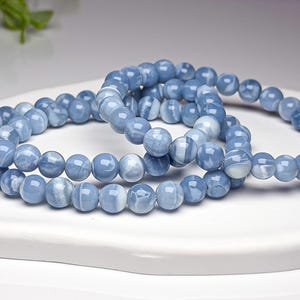 May include: A set of two stretch bracelets composed of smooth, round, light blue beads with subtle white marbled patterns. The beads have a polished, glossy finish and are strung together in a circular loop, creating a uniform appearance.