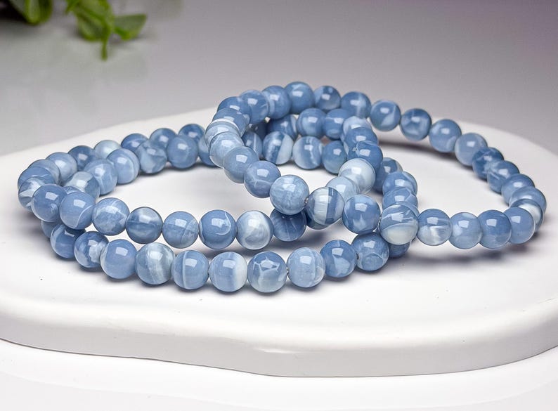 May include: A set of two stretch bracelets composed of smooth, round, light blue beads with subtle white marbled patterns. The beads have a polished, glossy finish and are strung together in a circular loop, creating a uniform appearance.