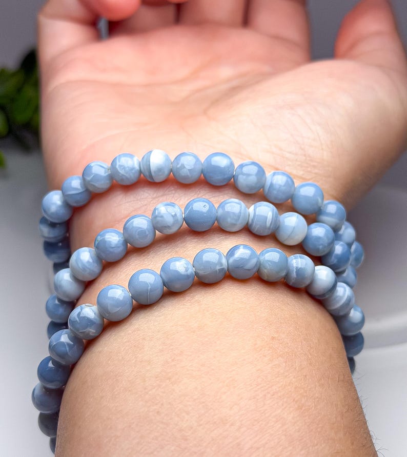 May include: A multi-strand bracelet composed of smooth, round, light blue beads with subtle white marbled patterns. The beads are strung together in a continuous loop, creating a layered appearance when wrapped around the wrist multiple times.