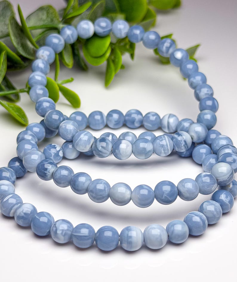 May include: A stretch bracelet composed of smooth, round, light blue beads with subtle white marbled patterns. The beads are uniform in size and arranged in a circular loop, showcasing a polished, reflective surface throughout the entire piece.