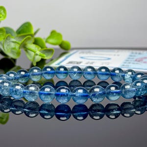 May include: A bracelet made of translucent, round, light blue beads. The beads are strung together, forming a complete circle. The bracelet is displayed on a reflective surface, with green leaves in the background.