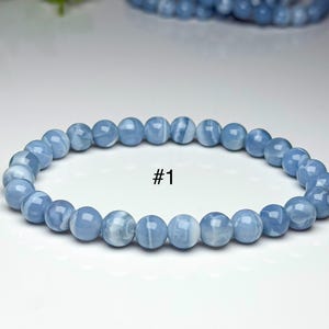 May include: A stretch bracelet composed of smooth, round, light blue beads with subtle white marbling throughout. The beads are strung in a single circular strand. The text #1 is printed in black in the center of the bracelet's open space.