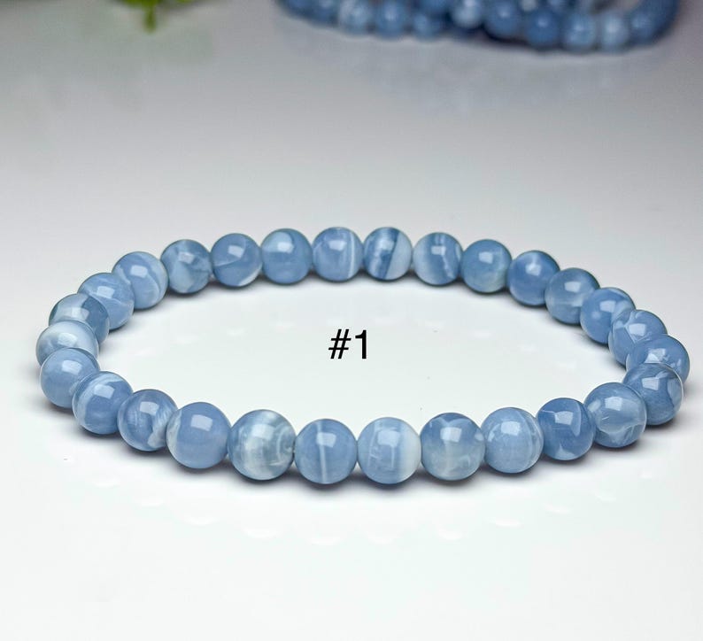 May include: A stretch bracelet composed of smooth, round, light blue beads with subtle white marbling throughout. The beads are strung in a single circular strand. The text #1 is printed in black in the center of the bracelet's open space.