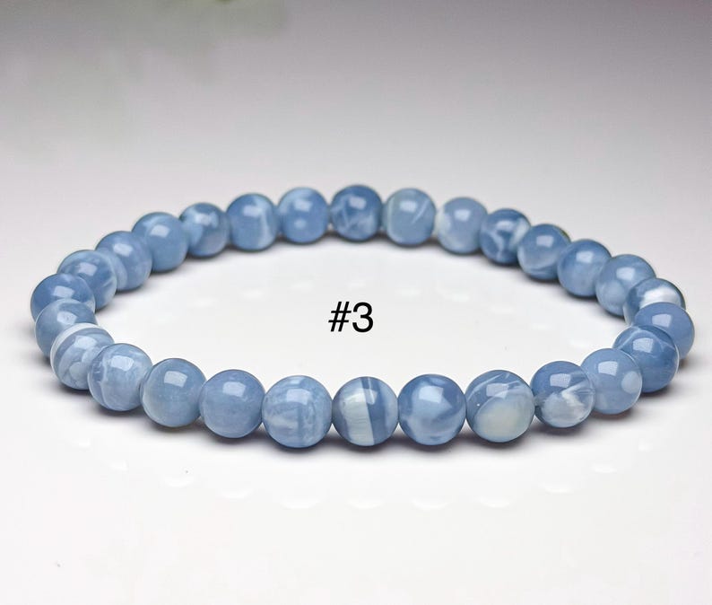 May include: A circular beaded bracelet featuring smooth, polished, light blue spherical beads with subtle white marbled patterns throughout. The bracelet is labeled with the text #3 in the center, showcasing a uniform arrangement of round stones.
