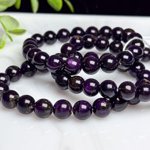 May include: A beaded bracelet featuring smooth, round, deep purple stones with a subtle reflective sheen. The beads are strung together on a flexible cord, creating a circular shape with a polished, glossy finish that catches the light.