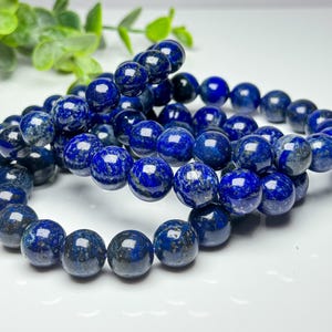 May include: A beaded bracelet composed of round, polished lapis lazuli stones in deep shades of blue with visible flecks of gold pyrite. The beads are strung together in a circular formation, showcasing a smooth, reflective surface texture.