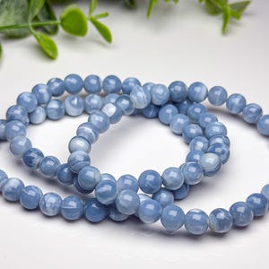May include: Two stretch bracelets composed of smooth, round, light blue beads with subtle white marbling patterns. The beads have a polished finish and are strung together in a circular shape, creating a consistent, uniform appearance throughout.