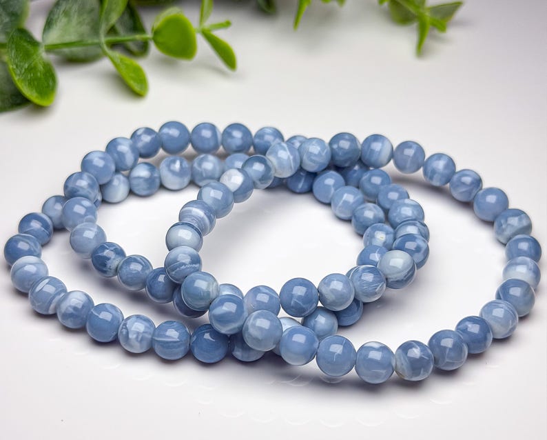 May include: Two stretch bracelets composed of smooth, round, light blue beads with subtle white marbling patterns. The beads have a polished finish and are strung together in a circular shape, creating a consistent, uniform appearance throughout.